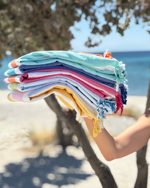 Stack of colorful Towel to Go Hammam towels held by a person on a beach with ocean and trees in the background