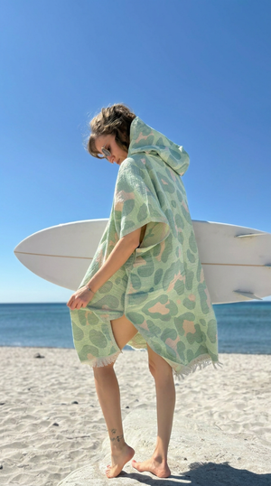 Women on a beach wearing a green patterned Towel to Go Jaquard Poncho with a surfboard.